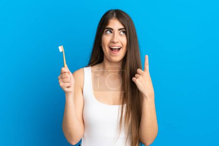 Photo for Young caucasian woman brushing teeth isolated on blue background thinking an idea pointing the finger up - Royalty Free Image