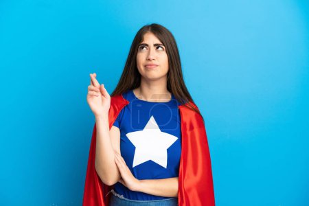 Téléchargez les photos : Super Hero femme caucasienne isolée sur fond bleu avec les doigts croisés et souhaitant le meilleur - en image libre de droit