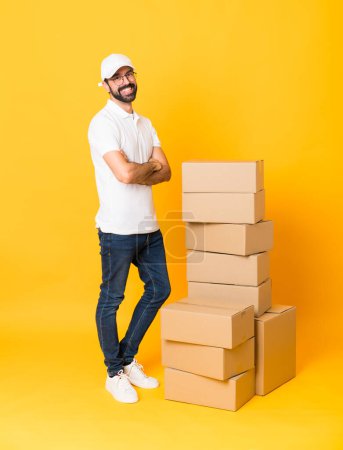 Téléchargez les photos : Plan complet de l'homme de livraison parmi les boîtes sur fond jaune isolé avec des lunettes et souriant - en image libre de droit