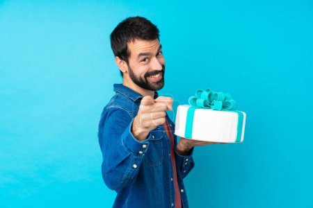 Foto de Joven hombre guapo con un gran pastel sobre fondo azul aislado señala el dedo en usted mientras sonríe - Imagen libre de derechos