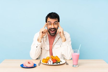 Téléchargez les photos : Homme à une table ayant des gaufres petit déjeuner et un milkshake avec des lunettes et surpris - en image libre de droit