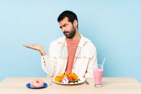 Téléchargez les photos : Homme à une table ayant des gaufres petit déjeuner et un milk-shake tenant copyspace avec des doutes - en image libre de droit