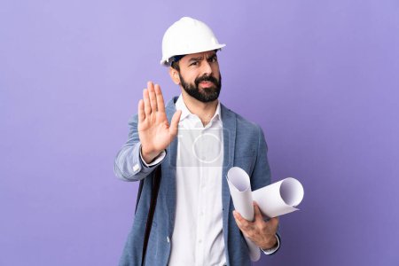 Téléchargez les photos : Jeune architecte homme avec casque et tenant des plans sur fond violet isolé faisant geste d'arrêt - en image libre de droit