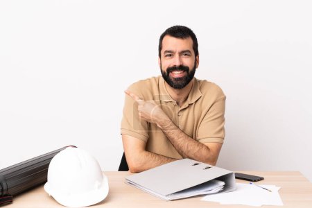 Foto de Arquitecto caucásico con barba en una mesa apuntando hacia un lado para presentar un producto. - Imagen libre de derechos