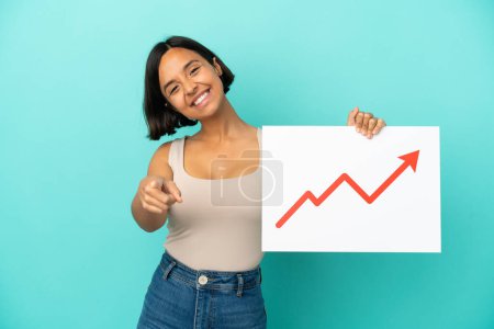 Photo for Young mixed race woman isolated on blue background holding a sign with a growing statistics arrow symbol and pointing to the front - Royalty Free Image