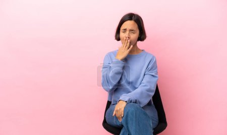 Foto de Mujer joven de raza mixta sentada en una silla aislada sobre fondo rosa bostezando y cubriendo la boca abierta con la mano - Imagen libre de derechos
