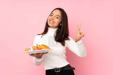 Young brunette woman holding waffles over isolated pink background showing victory sign with both hands