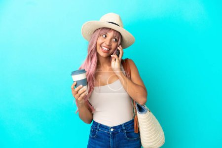 Foto de Joven mujer de raza mixta sosteniendo una bolsa de playa con pamela aislada sobre fondo azul sosteniendo café para llevar y un móvil - Imagen libre de derechos