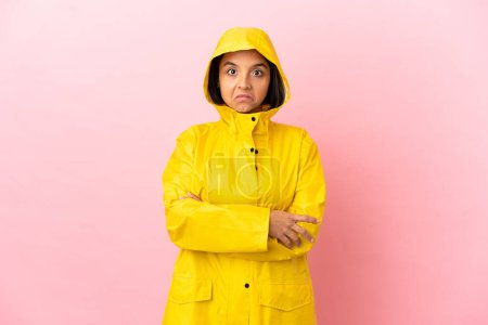 Junge lateinamerikanische Frau trägt einen regenfesten Mantel über isoliertem Hintergrund und macht eine Geste des Zweifels, während sie die Schultern hebt