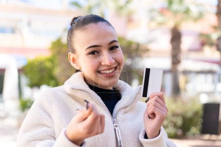 Téléchargez les photos : Jeune fille marocaine à l'extérieur tenant une carte de crédit faisant geste d'argent - en image libre de droit