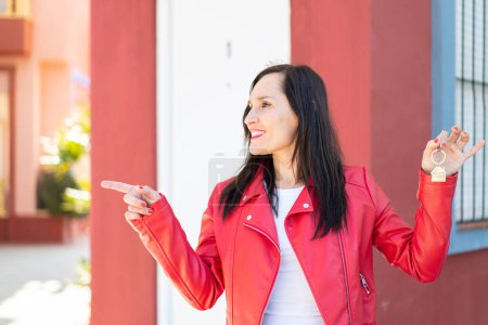 Téléchargez les photos : Femme d'âge moyen tenant les clés de la maison à l'extérieur pointant vers le côté pour présenter un produit - en image libre de droit