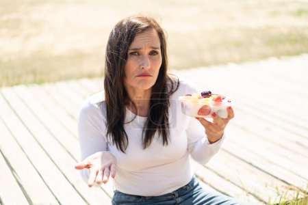 Photo for Middle aged woman holding a bowl of fruit at outdoors making doubts gesture while lifting the shoulders - Royalty Free Image