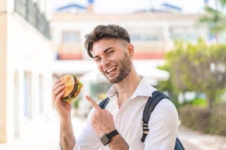Photo for Young handsome man holding a burger at outdoors and pointing it - Royalty Free Image