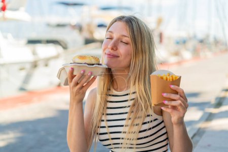 Photo for Young blonde woman at outdoors holding a burger and fried chips with happy expression - Royalty Free Image