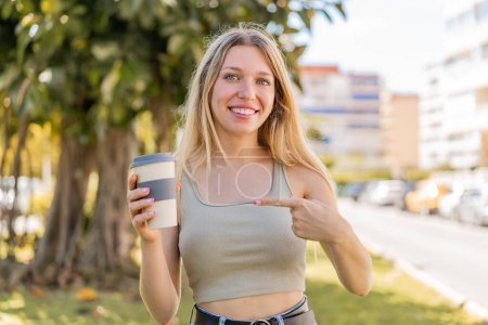 Young blonde woman holding a take away coffee at outdoors with surprise facial expression