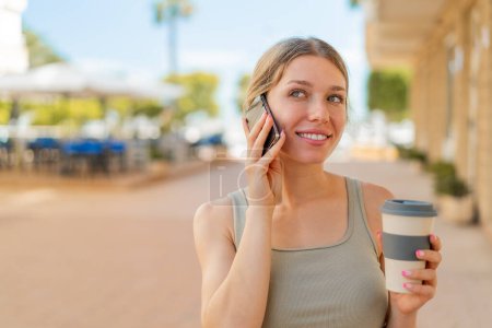 Photo for Young blonde woman at outdoors using mobile phone and holding a coffee - Royalty Free Image