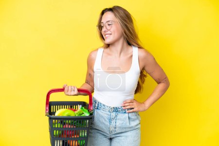 Photo for Young caucasian woman holding a shopping basket full of food isolated on yellow background posing with arms at hip and smiling - Royalty Free Image