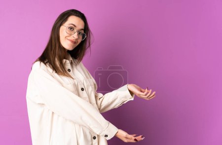 Téléchargez les photos : Jeune femme sur fond violet isolé étendant les mains sur le côté pour inviter à venir - en image libre de droit