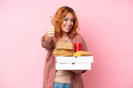 Foto de Joven mujer caucásica sosteniendo comida rápida aislada sobre fondo rosa con pulgares hacia arriba porque algo bueno ha sucedido - Imagen libre de derechos