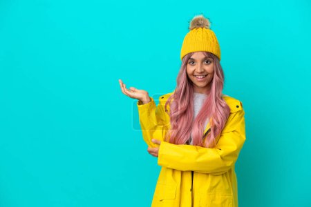 Young woman with pink hair wearing a rainproof coat isolated on blue background extending hands to the side for inviting to come