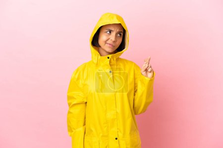Junge lateinische Frau trägt einen regendichten Mantel über isoliertem Hintergrund mit Daumendrücken und wünscht das Beste