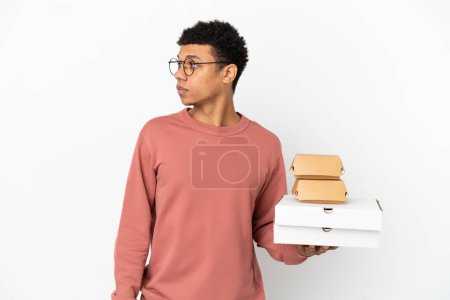 Téléchargez les photos : Jeune homme afro-américain tenant un hamburger et des pizzas isolé sur fond blanc regardant sur le côté - en image libre de droit