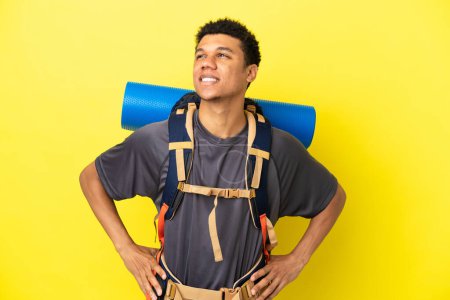 Téléchargez les photos : Jeune alpiniste afro-américain avec un gros sac à dos isolé sur fond jaune penser une idée tout en levant les yeux - en image libre de droit
