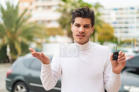 Junger Kaukasier, der Autoschlüssel im Freien hält, macht Zweifel, während er die Schultern hebt