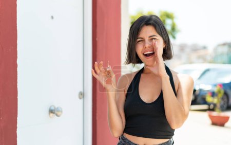 Foto de Joven bonita mujer búlgara sosteniendo las llaves de casa al aire libre gritando con la boca abierta - Imagen libre de derechos