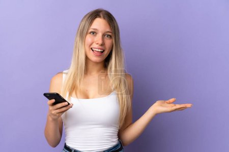 Téléchargez les photos : Jeune femme blonde utilisant un téléphone mobile isolé sur fond violet étendant les mains sur le côté pour inviter à venir - en image libre de droit
