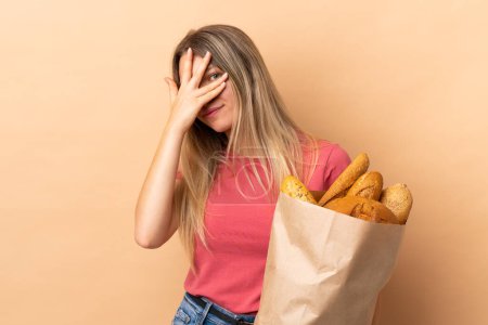 Young blonde woman holding a bag full of breads isolated on beige background covering eyes and looking through fingers