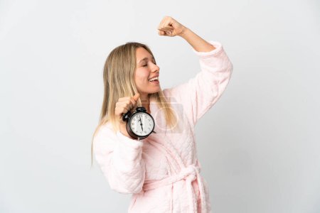 Téléchargez les photos : Jeune femme blonde isolée sur fond blanc en pyjama et tenant horloge faisant geste de victoire - en image libre de droit
