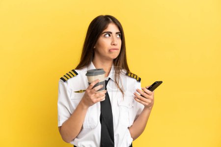 Airplane pilot isolated on yellow background holding coffee to take away and a mobile while thinking something