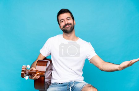 Junger Mann mit Gitarre vor isoliertem blauen Hintergrund präsentiert und lädt ein, mit der Hand zu kommen