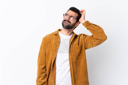 Foto de Caucásico hombre guapo con barba usando una chaqueta de pana sobre fondo blanco aislado que tiene dudas mientras se rasca la cabeza - Imagen libre de derechos