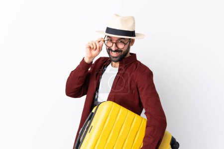 Foto de Hombre viajero con barba sosteniendo una maleta sobre un fondo blanco aislado con gafas y sonriendo - Imagen libre de derechos