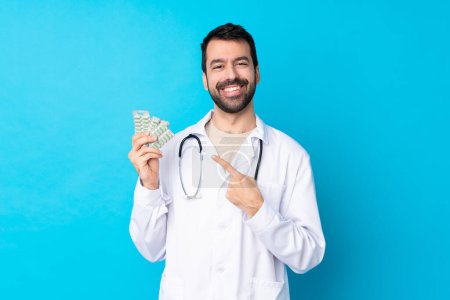Photo for Young caucasian man over isolated background wearing a doctor gown and holding pills - Royalty Free Image