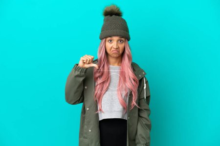 Young woman with pink hair wearing a rainproof coat isolated on blue background showing thumb down with negative expression
