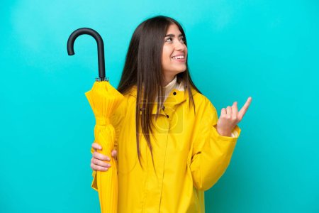 Young caucasian woman with rainproof coat and umbrella isolated on blue background pointing up a great idea