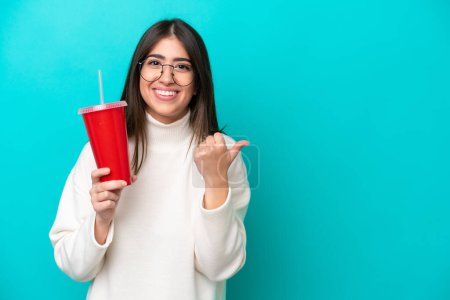 Téléchargez les photos : Jeune femme caucasienne buvant soda isolé sur fond bleu pointant vers le côté pour présenter un produit - en image libre de droit