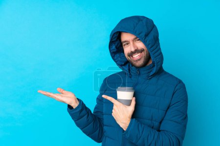Téléchargez les photos : Homme vêtu d'une veste d'hiver et tenant un café à emporter sur fond bleu isolé tenant un copyspace imaginaire sur la paume pour insérer une annonce - en image libre de droit