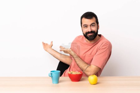Foto de Caucásico hombre desayunando en una mesa extendiendo las manos a un lado para invitar a venir. - Imagen libre de derechos