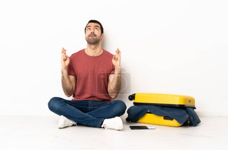 Photo for Caucasian handsome man with a suitcase full of clothes sitting on the floor at indoors with fingers crossing and wishing the best - Royalty Free Image