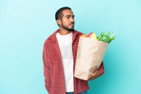 Foto de Joven latino sosteniendo una bolsa de compras aislada sobre fondo azul mirando a un lado - Imagen libre de derechos