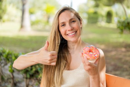 Téléchargez les photos : Jeune femme blonde tenant un beignet à l'extérieur avec les pouces levés parce que quelque chose de bon est arrivé - en image libre de droit