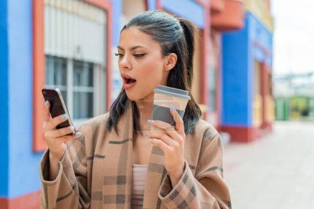 Téléchargez les photos : Jeune jolie femme à l'extérieur en utilisant un téléphone portable et tenant un café avec une expression surprise - en image libre de droit