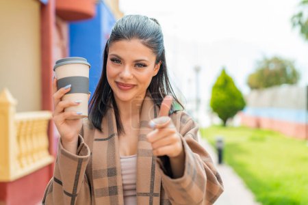 Téléchargez les photos : Jeune jolie femme tenant un café à emporter à l'extérieur pointe du doigt vers vous avec une expression confiante - en image libre de droit