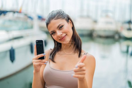 Téléchargez les photos : Jeune jolie femme utilisant un téléphone mobile à l'extérieur pointe du doigt vers vous avec une expression confiante - en image libre de droit
