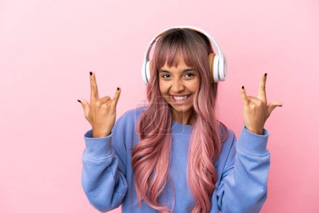 Foto de Joven mujer de raza mixta con cabello rosa aislado sobre fondo rosa escuchando música haciendo gesto rock - Imagen libre de derechos