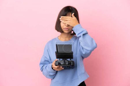 Young mixed race woman holding a drone remote control isolated on pink background covering eyes by hands. Do not want to see something